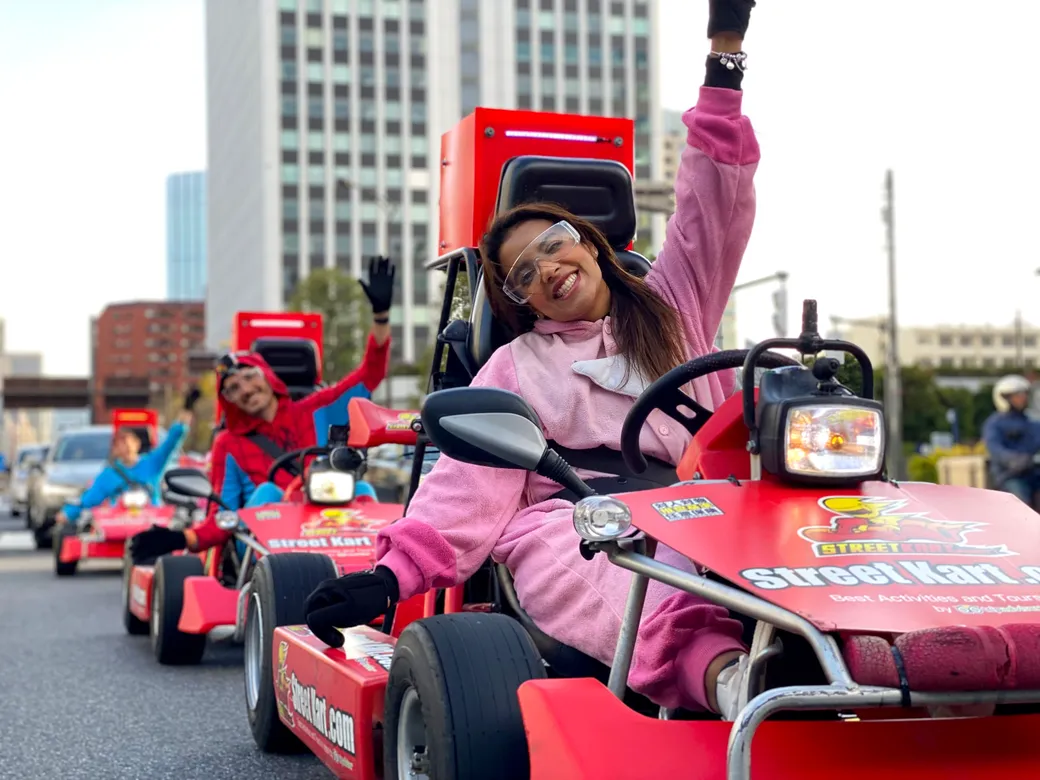 Smiling woman in a pink hoodie drives a red mini go-kart in a city street parade, raising her arm with others in similar karts behind her.