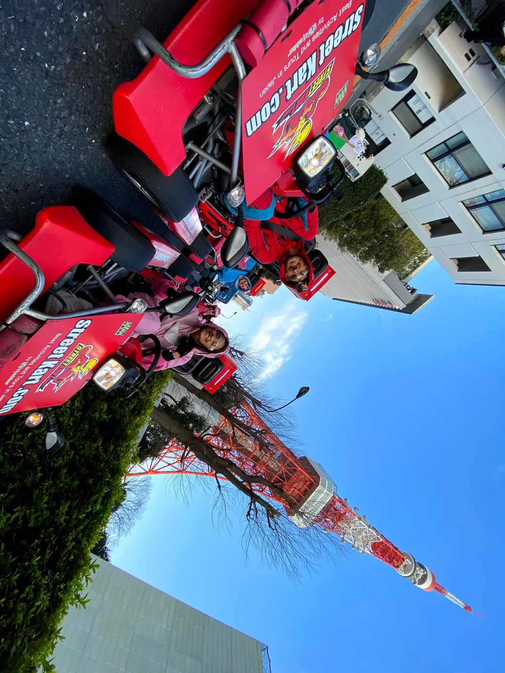 Line of red branded go-karts with people in red jackets smiling, banners readings promotional text, street parade scene with a tall red-and-white tower and clear blue sky in the background.