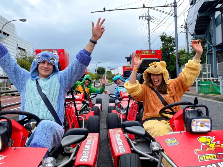 Group of people in plush animal costumes riding red go-karts in an urban street, waving at the camera with big smiles.